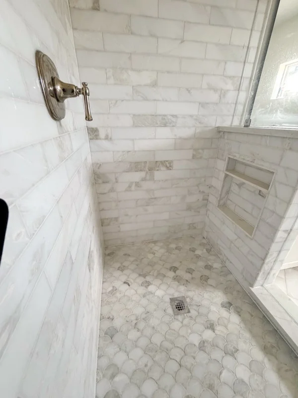 Walk-in shower with white marble subway tile walls, recessed tile niche, chrome valve, and gray and white fan-shaped mosaic floor tile
