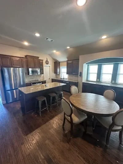 Modern kitchen with dark cabinets, stainless steel refrigerator, island, and adjacent dining area with hardwood flooring