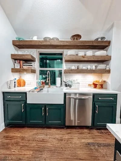 Kitchen with dark green cabinets, white sink, stainless steel dishwasher, wood floating shelves, subway tile backsplash