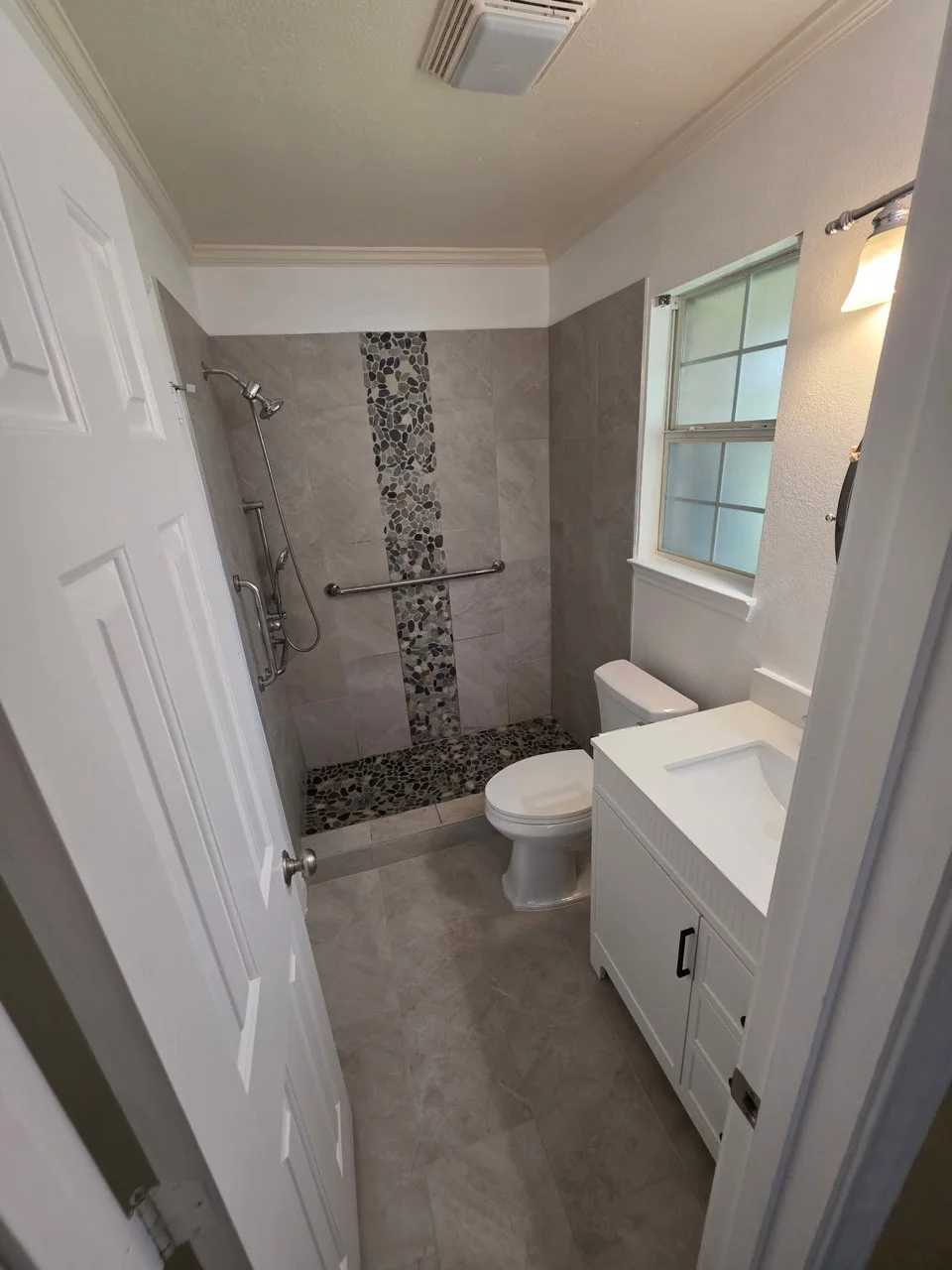 Walk-in shower with gray wall tile, pebble mosaic accent strip and shower floor, grab bar, and white vanity with quartz countertop