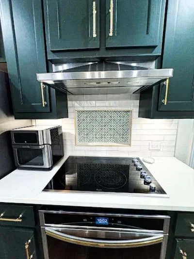 Dark teal cabinets, white quartz counters, geometric tile backsplash, stainless steel range hood and appliances