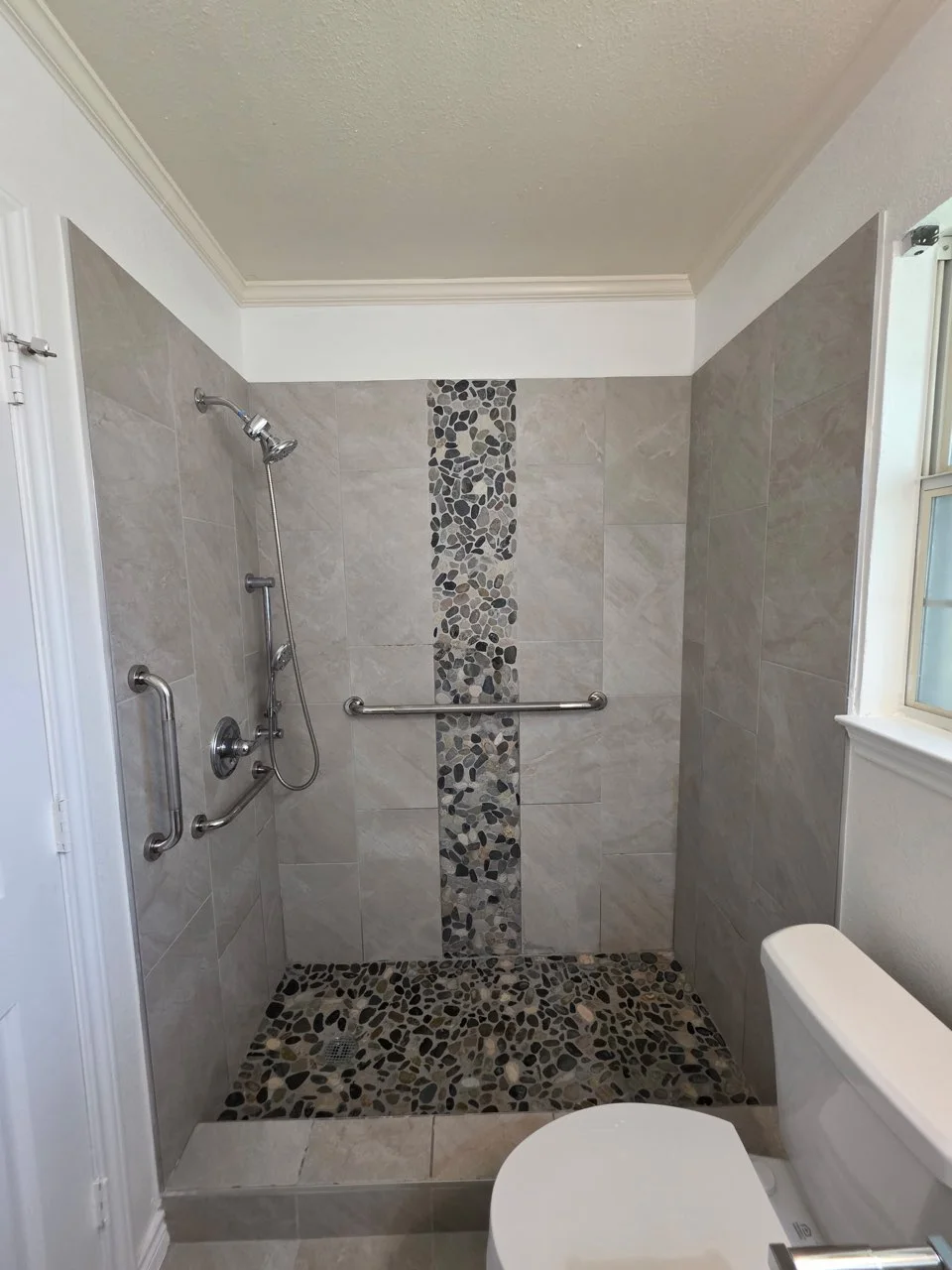 Walk-in shower with gray large-format wall tile, river pebble mosaic accent strip and floor, and chrome grab bars
